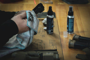 Person cleaning a handgun slide with a cloth on a Cerus Gear mat, surrounded by cleaning supplies and magazines.